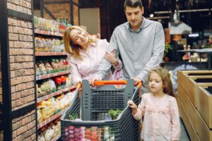 A family of three shopping in a supermarket, pushing a cart full of groceries.